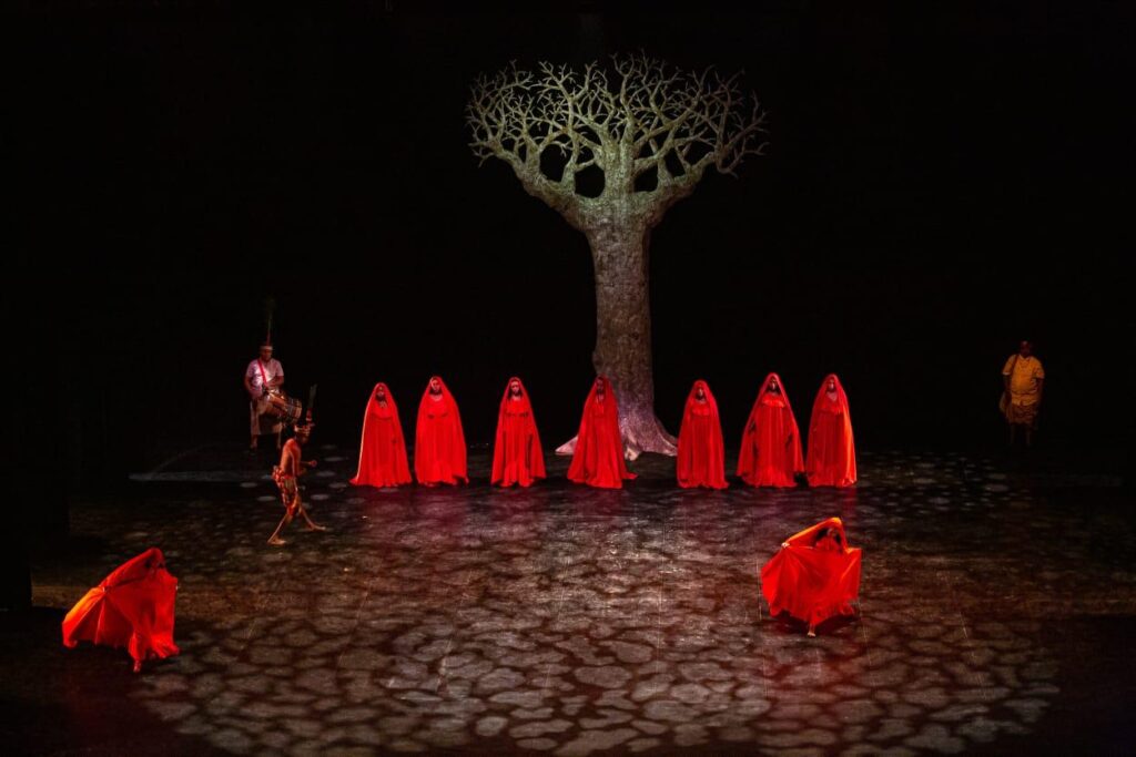 Performance se varias personas vestidas de rojo en un escenario que representa la tierra árida, de fondo se ve un árbol seco, durante una obra de teatro de la Agenda Cultural de la COP16.